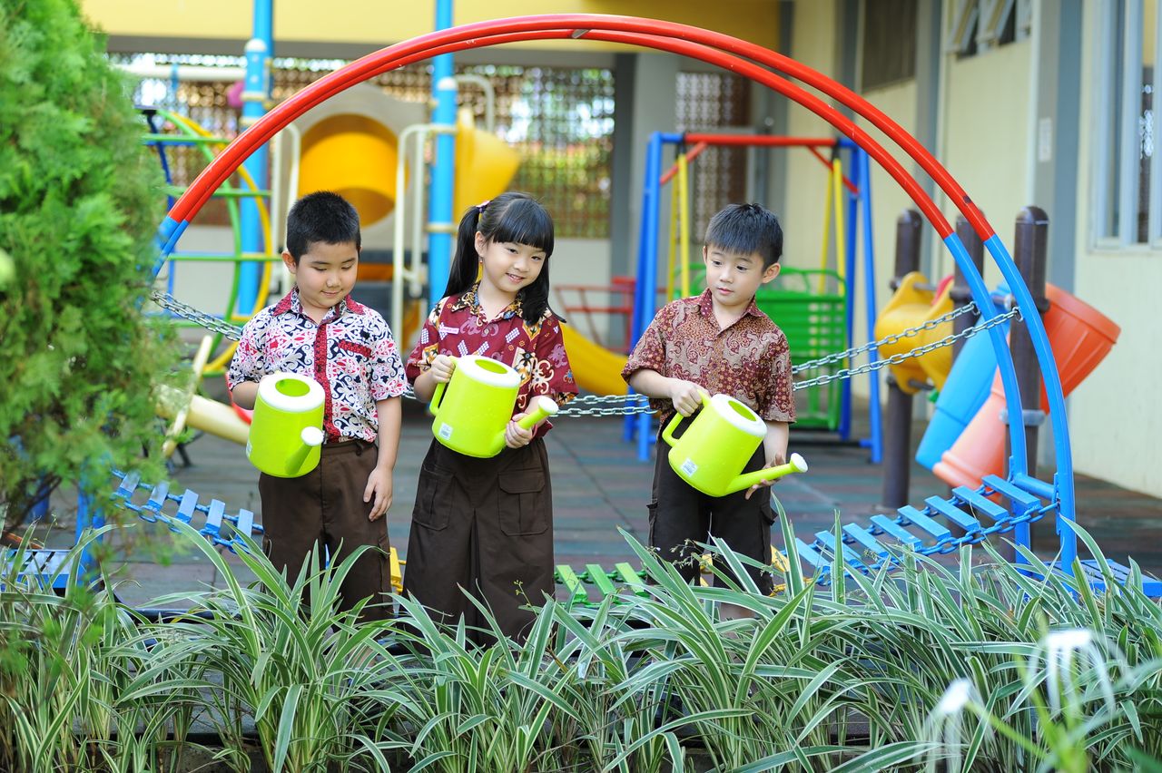 Group of children gardening at a playground with colorful play equipment and watering cans.
