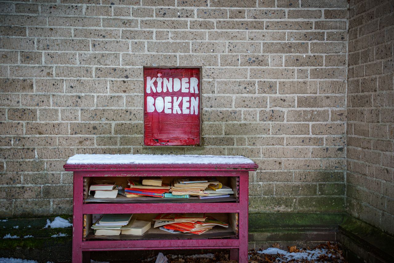 contact-img Red shelf with children's books marked 'Kinder Boeken' against a brick wall in winter.